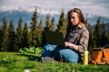 A young, slender girl with loose hair in a plaid shirt and jeans with an orange backpack works on a laptop sitting on green grass against the backdrop of mountains.