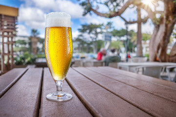 Cooled glass of beer on the wooden table. Blurred summer street cafe outside at the background. - obrazy, fototapety, plakaty
