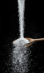 White refined sugar crystals falling down into the wooden spoon at black background.