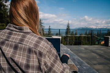 Fototapeta na wymiar a young, beautiful slender girl with flowing hair in a plaid shirt sits at an old wooden table and works at a laptop against the backdrop of mountains. View from the back.