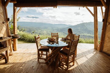 A young girl has breakfast in a bamboo house overlooking the mountains. Fruits, kettle of tea or coffee on the table overlooking the mountains in Bali.