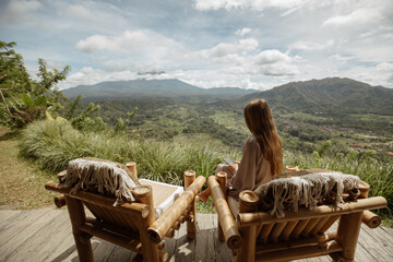 Young girl with a smartphone sits on a bamboo chair with a beautiful view of the mountains or the...