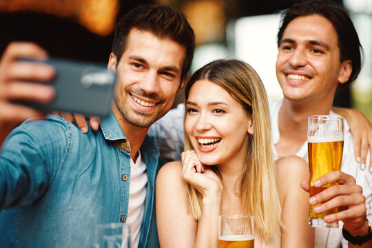 Group of young people have a good time in summer pub taking selfie at table and drink beer