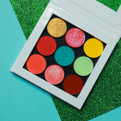 Rainbow spring and summer eyeshadow makeup palette. Realistic image with matte, glitter, multichrome pigments. Top view flat lay package isolated on green glitter and light blue background