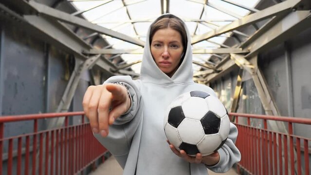 Young urban woman football player holding soccer ball pointing with finger to the camera and to you.