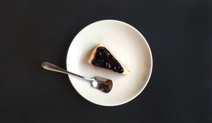 Top view of slice of blueberry cheesecake and stainless steel spoon in white dish or plate on black background with copy space. Flat lay of sweet dessert and Homemade food on dark table