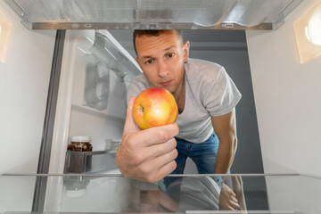 Man takes out a single apple from an empty refrigerator. Concept of delivery service, hunger, dients . Photo from inside the refrigerator - obrazy, fototapety, plakaty