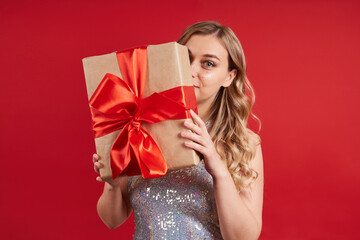 Charming young lady in a silvery dress hides behind a gift box, isolated on a red background