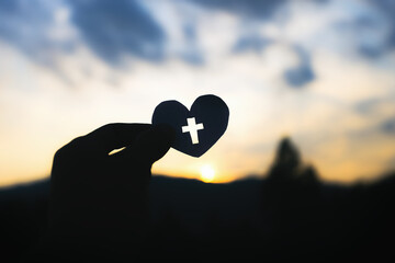 Hand holding cross in heart shapes. with light of sunset background, Symbol of Love, christian silhouette concept.