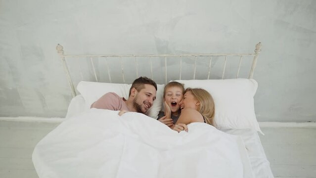 A Young Family Woke Up In The Morning In A White Bed With A Good Mood. Blonde Mom Hugs Her Son Lying In Bed.