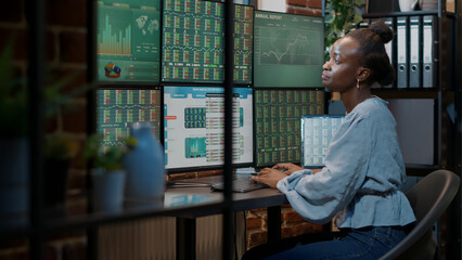 Bank employee analyzing forex exchange sales on laptop and multiple monitors in office, working to...