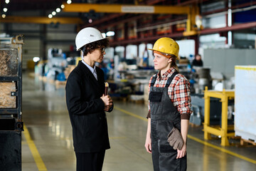 Young female engineer in workwear and inspection manager in black suit standing in large workshop of modern factory and communicating