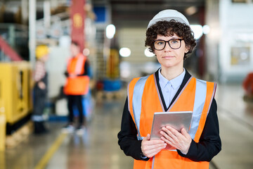 Young confident manager of factory in hardhat and uniform holding digital tablet while standing in front of camera in workshop