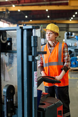 Female engineer looking forwards while standing in front of electric forklift and moving along...