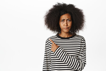 Close up of skeptical african female model frowning, looking displeased and pointing finger left, showing bad advertisement, standing over white background