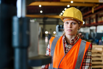Young female worker of modern factory in orange jacket and protective helmet looking at camera in large workshop or warehouse - obrazy, fototapety, plakaty