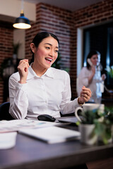 Happy joyful asian businesswoman celebrating successful startup project while sitting at desk. Enthusiastic excited office worker in workspace being positive about company profits.