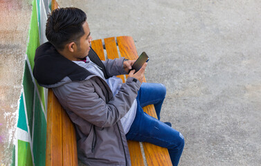 high angle shot of a mexican young man sitting on a bench of a square while using his smartphone