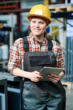 Young successful female engineer in coveralls and hardhat holding tablet while standing in front of camera against interior of warehouse