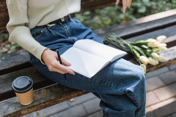 cropped view of woman in jeans holding notebook near bouquet of tulips and paper cup on bench.