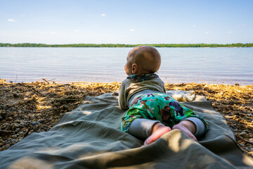 Aa attentive baby lies on his belly by a lake and looks into the distance