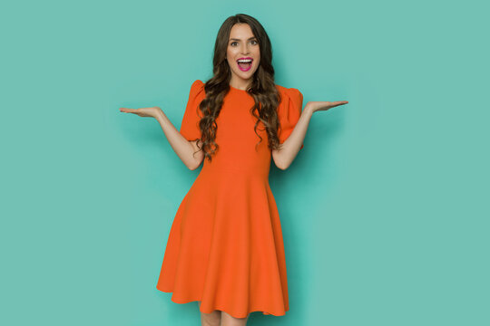 Happy young woman in orange mini dress is posing with hands raised and shouting.