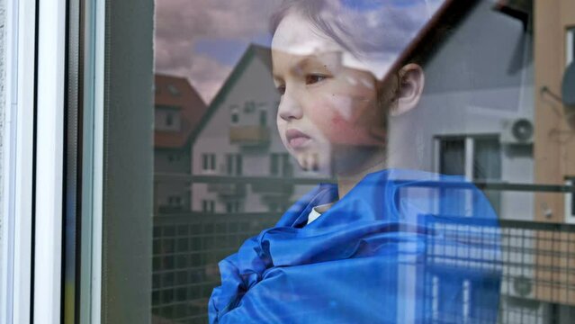 Little girl with the flag of Ukraine on her shoulders looks out the window with a serious expression.