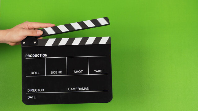 The hand is holding a black clapper board or movie slate on the green screen background.