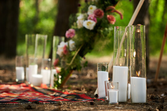 Candles burning in glass vases, flasks on carpet near bohemian tipi arch decorated with roses and flowers wrapped in fairy lights on outdoor wedding ceremony venue in pine forest. Boho rustic decor.