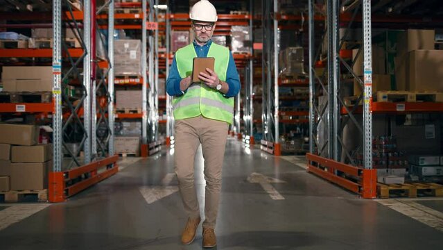 Concentrated handsome Caucasian professional male manager wearing helmet typing on tablet browsing online doing inventory walking in distribution center with many shelves and boxes. Warehouse job