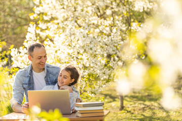 A girl and a young father are sitting at a laptop and studying in a flowered garden. Against the background of green grass and flowering trees. Remotely buy items in a store