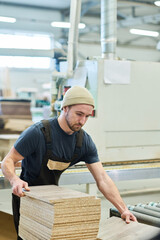 Young carpenter in uniform working with wooden boards at machine at furniture factory