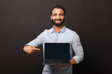 Happy Indian man holding laptop with empty screen and points finger at it, hispanic male employee...