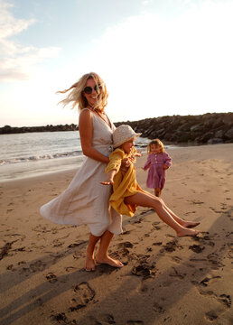 Joyful mother with two daughters playing on the beach, having fun during sunset, relaxing. Summer vacations with children. Concept of friendly family time on holidays.