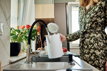 Woman pouring clean filtered water from faucet into electric kettle for boiling water at kitchen - Powered by Adobe