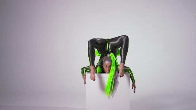 Front view confident flexible woman with futuristic makeup in costume looking at camera doing gymnastic split on cube upside down. Portrait of young slim Caucasian gymnast posing performing