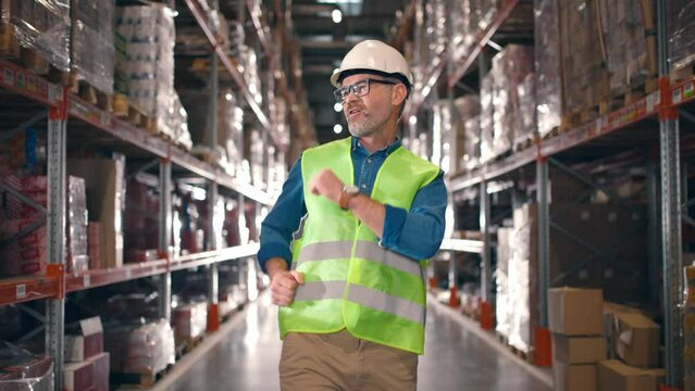 Happy positive Caucasian middle-aged man in helmet and eyeglasses walking in rows with many shelves with goods in boxes and dancing in good mood. Construction, factory, logistics center, work concept