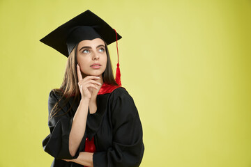 Front view of beautiful girl in mortarboard and graduate gown standing, thinking. Pretty brunette female looking up, asking, dreaming. Isolated on green studio background.