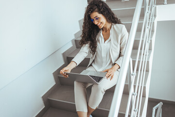 Beautiful young woman working on laptop and smiling while sitting at the staircase. Beautiful business woman sitting on the stairs in cafe on stairs and working with laptop on the legs.