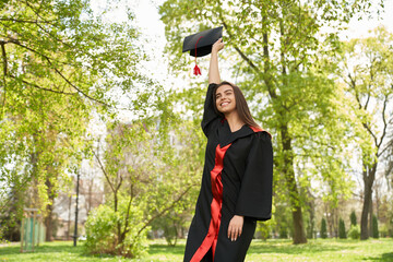 Front view of pretty girl wearing graduate gown raising mortarboard. Brunette young female graduating from college, university, standing with closed eyes. Concept of knowledge.