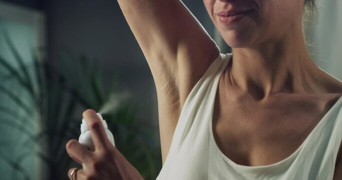 Cinematic macro of young woman in white tank top is applying female antiperspirant body spray deodorant on her armpit during morning beauty and personal hygiene routine at home before going out.