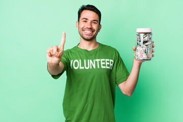 young adult hispanic man smiling and looking friendly, showing number one. donation concept