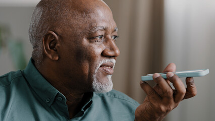 Mature grey-haired 60s biracial man with beard african american senior old male at home hold smartphone speak to phone microphone speaker audio voice message using speakerphone talking mobile app