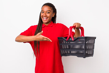 young adult black woman smiling cheerfully, feeling happy and showing a concept. empty shopping basket concept - obrazy, fototapety, plakaty