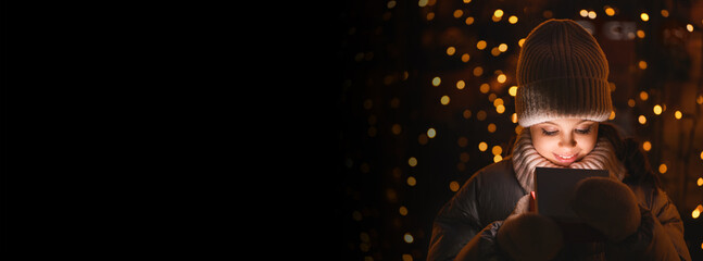 Girl and gift box with Christmas lights. Smiling girl in a hat, scarf looking into a glowing box on black background with blurry lights, copy space. Banner