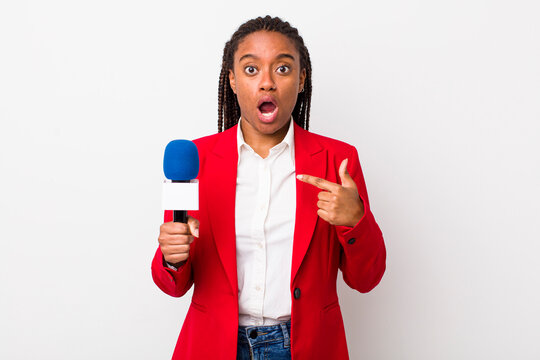 young adult black woman looking shocked and surprised with mouth wide open, pointing to self. presenter and microphone concept