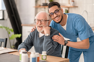 Merry male nurse hugging a pensioner in the kitchen - obrazy, fototapety, plakaty