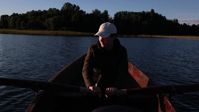 Young man is rowing with wooden oars on wooden boat on a lake during sunny day