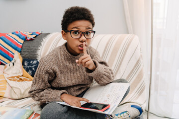 Black boy gesturing while using mobile phone and doing homework on sofa