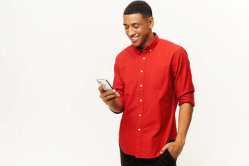 Smiling optimistic African-American young man in red casual shirt using smartphone isolated on...